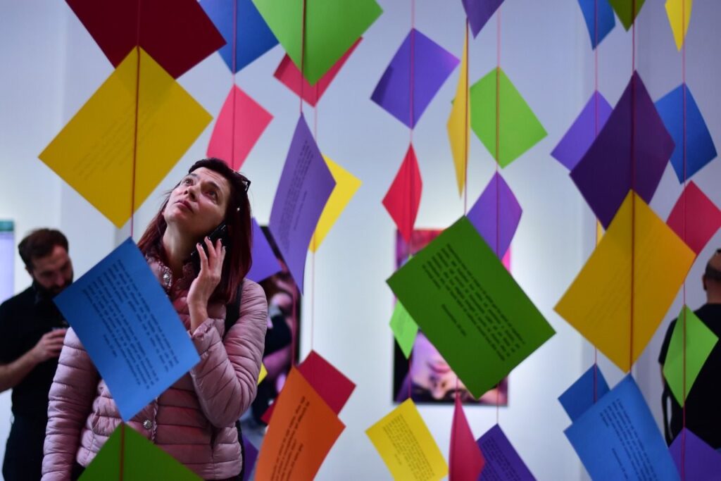 Person with audio device stands amid colored paper suspended from ceiling by red yarn
